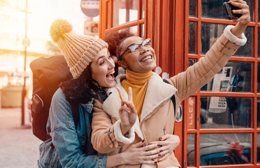 two friend, girlfriend and women using a mobile phone, camera and taking selfie against a red phonebox in the city of England.Travel Lifestyle concept