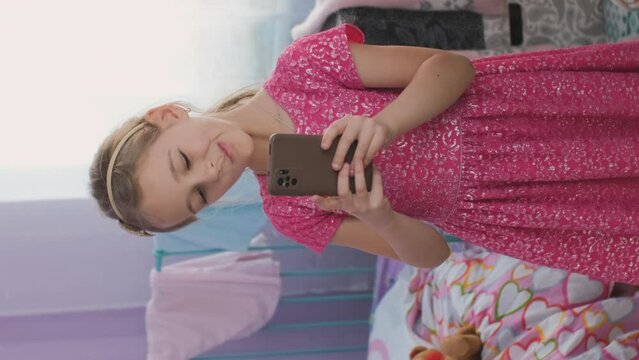 Vertical video. Little girl in pink dress taking selfies on her smartphone in messy room, grimacing and having fun at home
