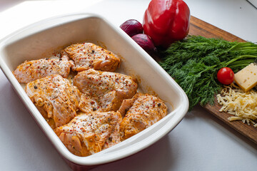 Raw marinated chicken legs in ceramic cooking tray. Fresh vegetables on cutting board
