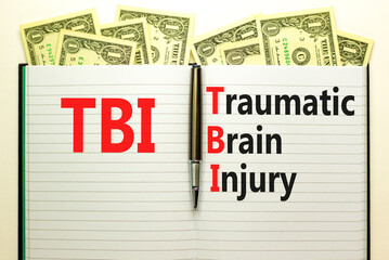 TBI traumatic brain injury symbol. Concept words TBI traumatic brain injury on white note on a beautiful background from dollar bills. Pen. Medical and TBI traumatic brain injury concept. Copy space