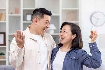 A young Asian couple, a man and a woman, stand hugging in a house, apartment, and hold keys in their hands. Smiling people look at each other, happy buyers, rent.