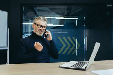 Angry senior businessman sitting in the office at the table with a laptop and worriedly talking on the phone, clenching his fist.