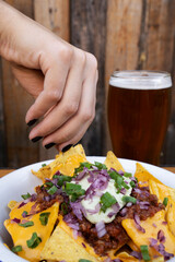 Eating nachos. Closeup view of a woman's hand holding a nacho dipped in cheddar cheese. A glass of beer besides the white bowl.