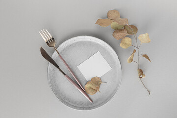 Festive table setting. Golden modern cutlery, dried eucalyptus branch, ceramic plate and blank paper as mock up for invitation or congratulation on white background