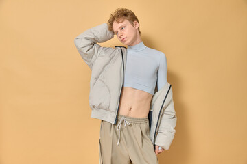 Stylish thoughtful serious handsome male with red curly hair wearing female clothes grey crop top posing with his arm bent in elbow behind his head on yellow background copy space. Queer fashion.