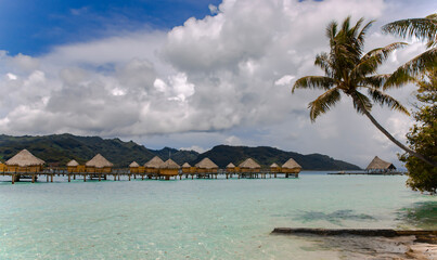 Overwater bungalows stretching out across the lagoon and a few catamarans in a quiet bay in Bora Bora island, Tahiti. Romantic honeymoon destination.