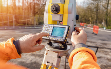 Site engineer operating his instrument during roadworks. Builder using total positioning station tachymeter on construction site for new road setting out - obrazy, fototapety, plakaty