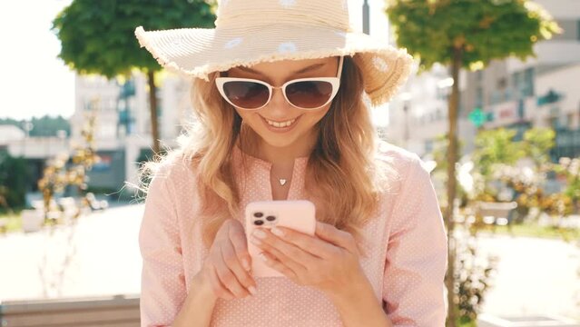 Young beautiful smiling hipster woman in trendy summer clothes. carefree female posing on the street background in sunglasses. Positive model outdoors using smartphone apps, looking at the screen