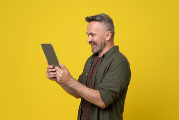 A playfully happy senior man dressed in green shirt looking into digital tablet pc gadget holding in hands reading news or watching video isolated on yellow background