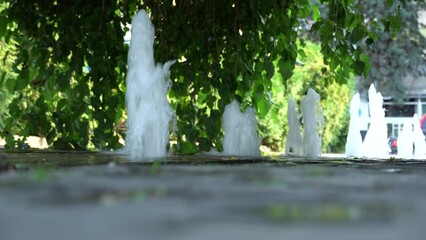Barefoot young woman walking in pedestrian fountain. Girl white t-shirt and green shorts go along of cascade water in city park. Refreshing cool on hot summer day. Person enjoy stream fountain.