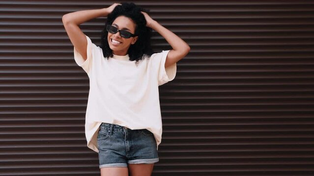 Beautiful black woman with afro curls hairstyle. Smiling hipster model in white t-shirt. Sexy carefree female posing in the street near roller shutter brown wall. Cheerful and happy, in sunglasses