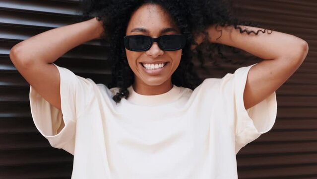 Beautiful black woman with afro curls hairstyle. Smiling hipster model in white t-shirt. Sexy carefree female posing in the street near roller shutter brown wall. Cheerful and happy, in sunglasses