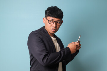 Portrait of young Asian business man in casual suit looking surprised at the camera while holding phone. Isolated image on blue background