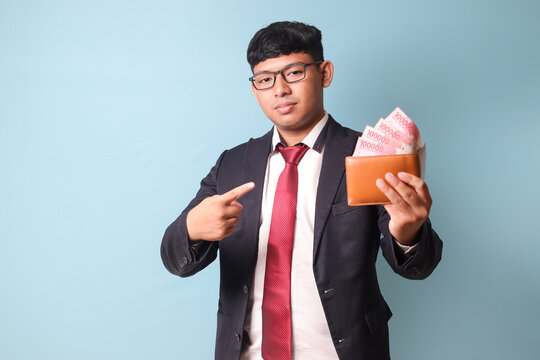 Portrait of young Asian business man in casual suit holding and pointing at leather wallet with thousand rupiahs. Isolated image on blue background