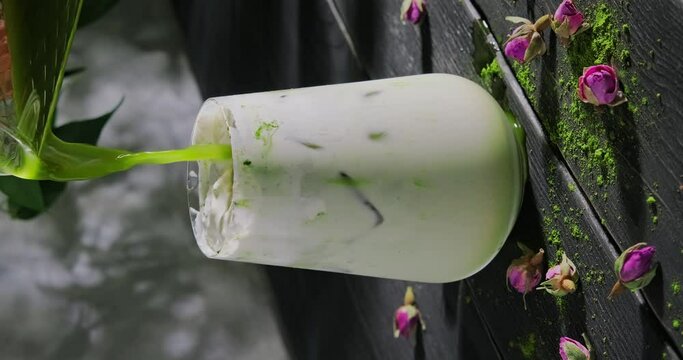 Preparing matcha latte with milk foam, pouring green tea in a glass with milk and ice cubes, cool refreshing drink with antioxidants, slow motion vertical video, high quality 4k footage