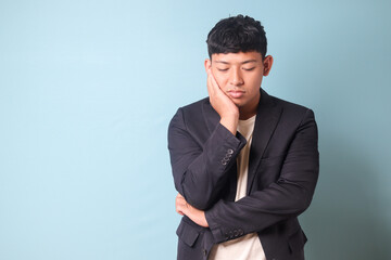 Portrait of young Asian business man in casual suit thingking hard with sleepy expression. Isolated image on blue background