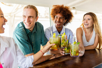 Multiracial young friends cheering with mojitos at terrace beach bar. Happy diverse people having fun drinking cocktails on the beach. Summer lifestyle and vacation concept