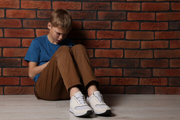 Upset boy sitting on floor near brick wall, space for text. Children's bullying