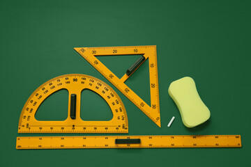 Protractor, triangle, ruler and sponge on green chalkboard, flat lay