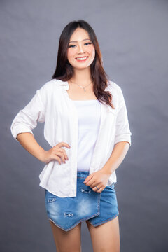 happy smiling asian woman posing standing in front of the camera holding her waist and hotpen indoors. fashion promotion women's shirt photo session in gray background studio