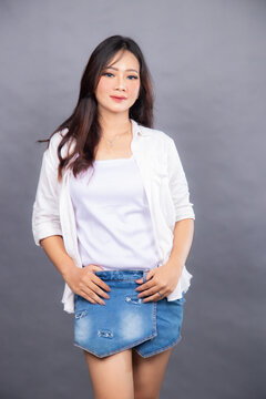 elegant asian woman posing standing in front of the camera while her hands holding hotpen jeans indoors. fashion promotion women's shirt photo session in gray background studio