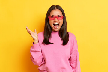 Angry black-haired woman in pink sunglasses and pink sweatshirt in the studio on a yellow...