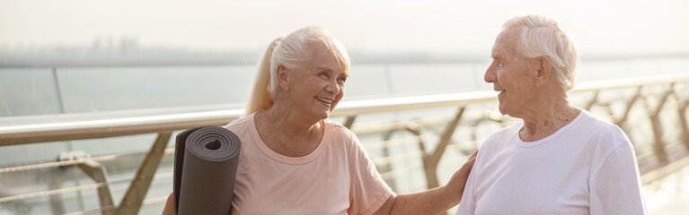 Smiling senior woman with mat cheers up man with poles for Nordic walking on footbridge