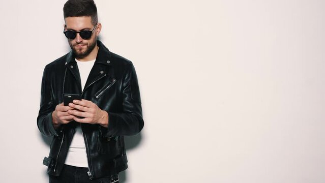 Handsome confident model. Sexy stylish man dressed in biker leather jacket and black jeans.Fashion hipster male in studio in sunglasses. Looking at smartphone screen. Using apps. Scrolling, texting