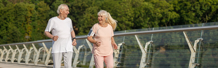 Happy senior man and woman run together along footbridge in summer
