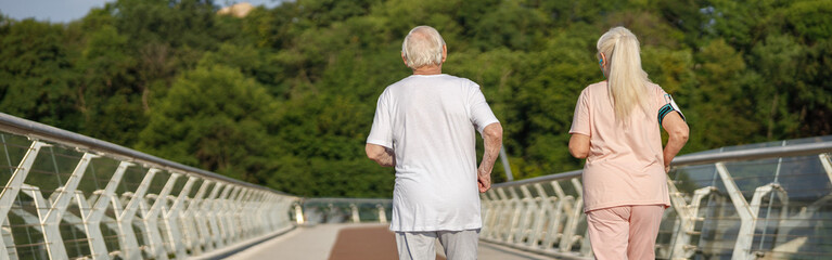 Sportive senior man and woman in tracksuits run together along footbridge