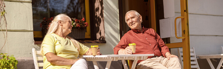 Positive mature man and woman with drinks rest at small table in cozy street cafe