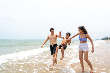 Happy Asian family travel ocean on summer holiday vacation. Parents and little son in swimwear have fun outdoor activity lifestyle walking and playing sea water together at tropical beach in sunny day