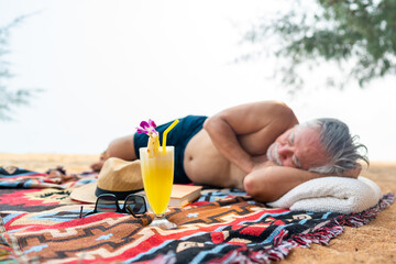 Asian senior man napping and sunbathing at tropical beach in sunny day. Retired elderly people relax and enjoy outdoor activity lifestyle travel nature ocean with family on summer holiday vacation.