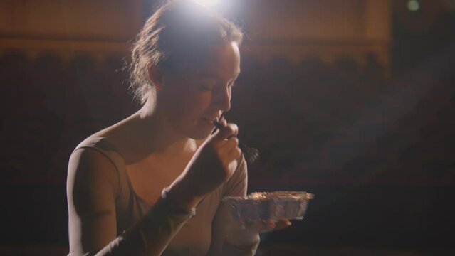 Beautiful ballerina eats and discusses performance with partner after choreography rehearsal. Ballet female dancer sits on theatre stage. Empty rows in theatrical hall. Ballet concept. Portrait.