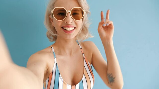 Young smiling blond model in summer swimwear colourful bathing suit. Sexy carefree woman having fun and going crazy. Female posing near blue wall in studio. Cheerful and happy in sunglasses.
