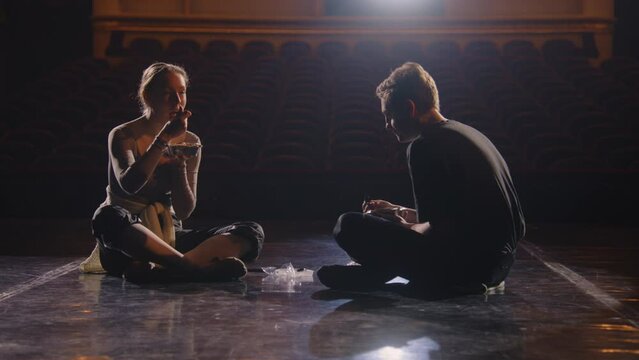 Pair of weary classical ballet dancers sit on theatre stage after choreography rehearsal. Ballerina eats with male dance partner after training. Empty rows in theatrical hall. Concept of ballet art.