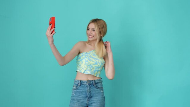Young beautiful smiling blond female in trendy summer clothes. Carefree woman posing near blue wall in studio. Positive model having fun indoors. Cheerful and happy. Taking selfie photos