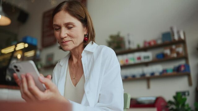 Cheerful elderly woman in white shirt typing on mobile in cafe