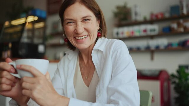 Happy elderly woman in white shirt drinking tea and looking at camera in cafe