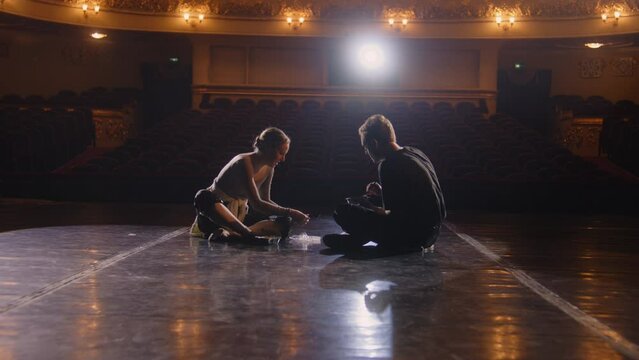 Pair of tired ballet dancers sit on theatre stage after choreography rehearsal. Ballerina eats with male dance partner after training. Empty rows in theatrical hall. Concept of classical ballet art.