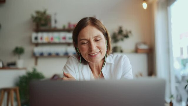 Happy old woman in white shirt talking by video call in headphones on laptop in cafe