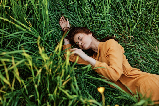 a close horizontal photo of a pleasant woman in a long orange dress resting lying in the tall grass with her eyes closed in sunny weather with her arms outstretched