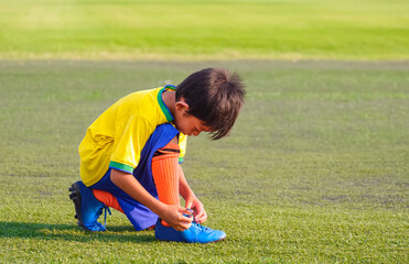 Fototapeta na wymiar Asian little boy in soccer uniform tying shoelace on soccer field before training
