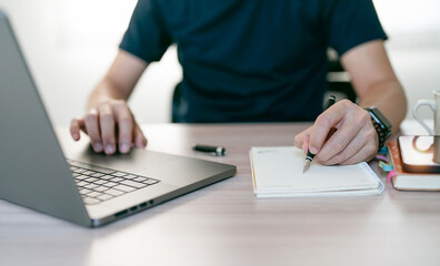 Young man writing down on notebook and using laptop computer, sitting at home office.