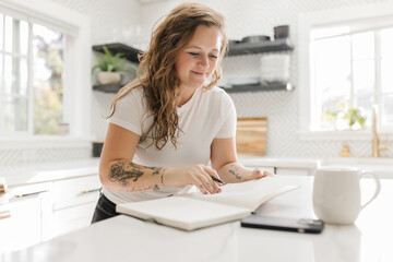 Happy Mid-Adult standing at kitchen counter journaling with coffee