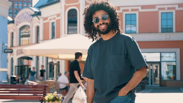 Handsome smiling hipster  model. Sexy unshaven Arabian man dressed in summer clothes. Fashion male with long curly hairstyle posing in the street at sunset. Cheerful and happy. In sunglasses 