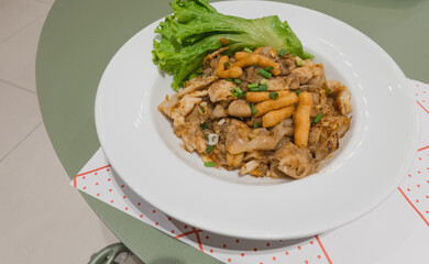 Kuaitiao khua kai or stir fried rice noodles with chicken topped with spring onion and crispy dough stick, lettuce, white plate, top view, green table, Thai dishes