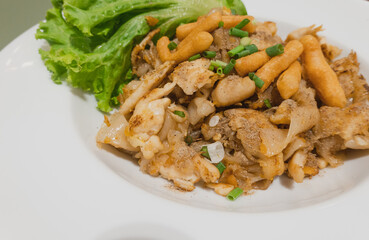 Kuaitiao khua kai or stir fried rice noodles with chicken topped with spring onion and crispy dough stick, lettuce, white plate, close up, Thai dishes