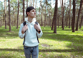 A young man in a white sweater and hat walks in the woods. Male tourist in nature. Man with a dark backpack and light jeans