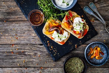 Tasty sandwiches - toasted bread with burrata cheese, anchovies, bell pepper, olives, tomatoes and thyme on wooden table
